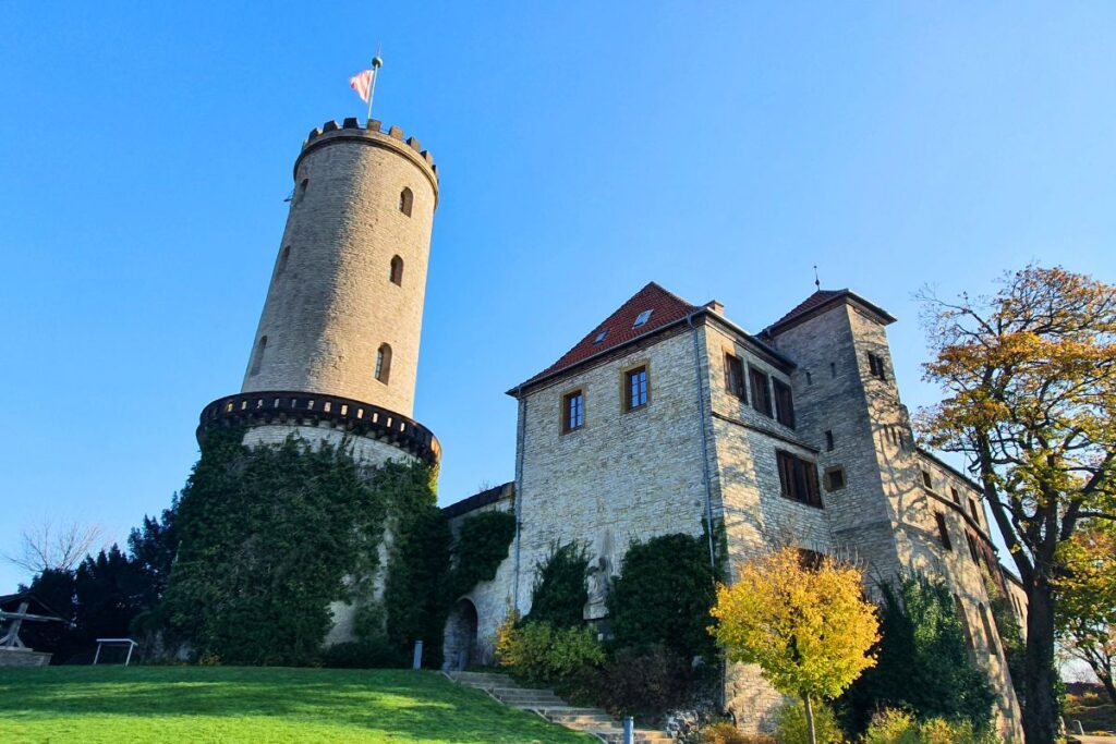 Der mächtige runde Festungsturm der Sparrenburg mit wehender Flagge über einer Wallanlage im Sonnenschein in Bielefeld.